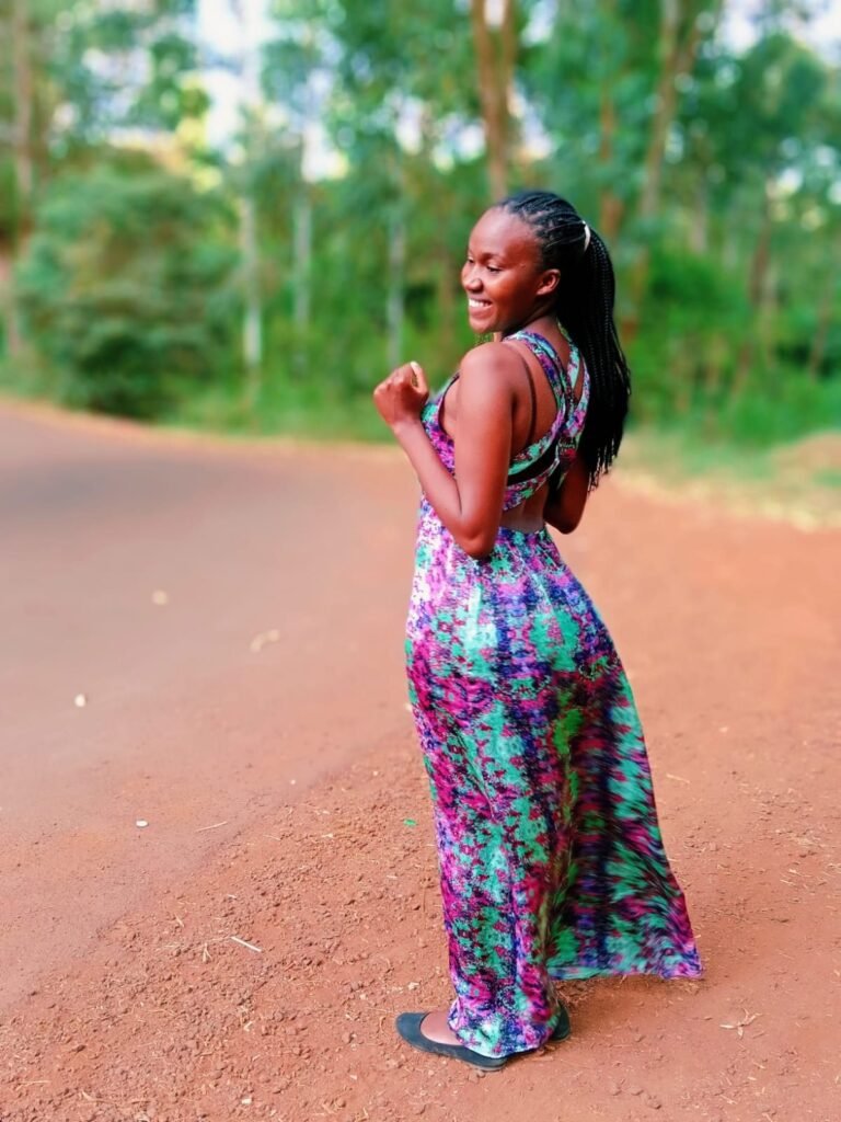 Christine Murugi Gitonga smiling on a red dirt road lined with trees, in a long green and purple dress
